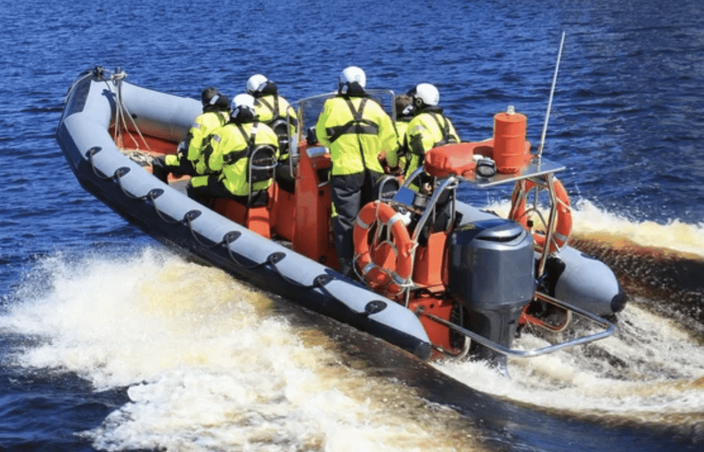 Screenshot 2026-03-15 at 000121 - N9BO℠ | Global Underwater Services Ltd A group of people in bright yellow jackets and helmets ride a grey inflatable motorboat across dark blue water, creating a foamy wake behind them. The boat is equipped with safety gear and an outboard motor.
