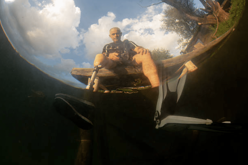 Screenshot 2026-03-15 at 103837 - N9BO℠ | Global Underwater Services Ltd A man with a prosthetic leg sits on a wooden jetty above water, preparing to dive, with one leg submerged and wearing a swim fin; trees and clouds are visible in the background.