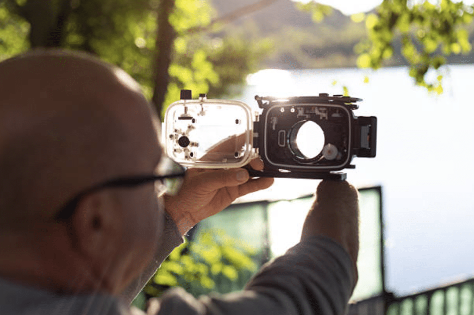 Screenshot 2026-03-15 at 103849 - N9BO℠ | Global Underwater Services Ltd A person holds up an open underwater camera housing towards a sunlit lake, with green trees and a metal fence visible in the background. The sunlight is shining through the housing’s opening.