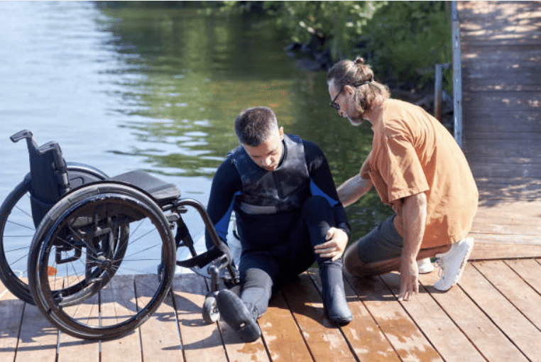 Screenshot 2026-03-15 at 103911 - N9BO℠ | Global Underwater Services Ltd A man in a wetsuit sits on a jetty next to a wheelchair, with another man kneeling beside him, offering support near a body of water and a ramp surrounded by greenery.