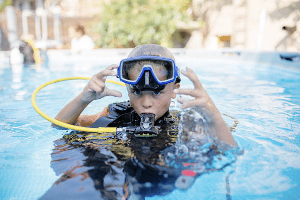 A young boy wearing scuba diving kit, including a mask and regulator, is in a swimming pool. He is raising his hands playfully near his head whilst looking at the camera; bubbles are visible in the water.