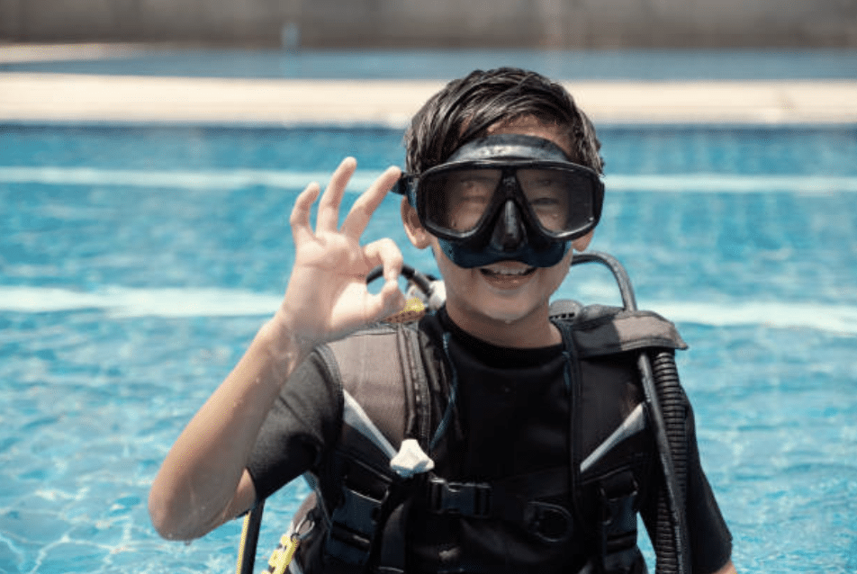A smiling young boy wearing scuba gear and a diving mask stands in a swimming pool, holding up his hand to make an OK sign, signalling that he is ready or feeling good.