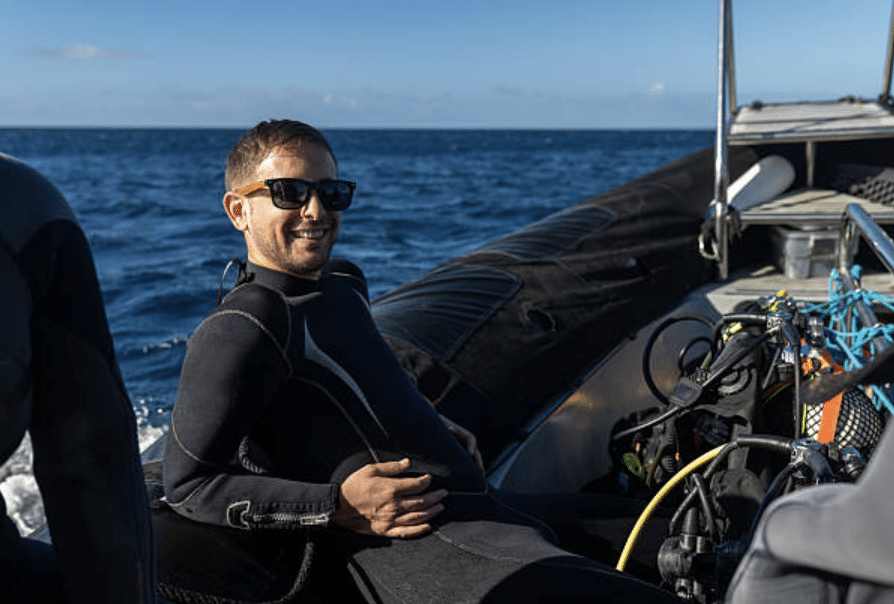Screenshot 2026-03-15 at 123954 - N9BO℠ | Global Underwater Services Ltd A man in a wetsuit and sunglasses sits on a boat with diving equipment, smiling, with the sea and blue sky in the background.