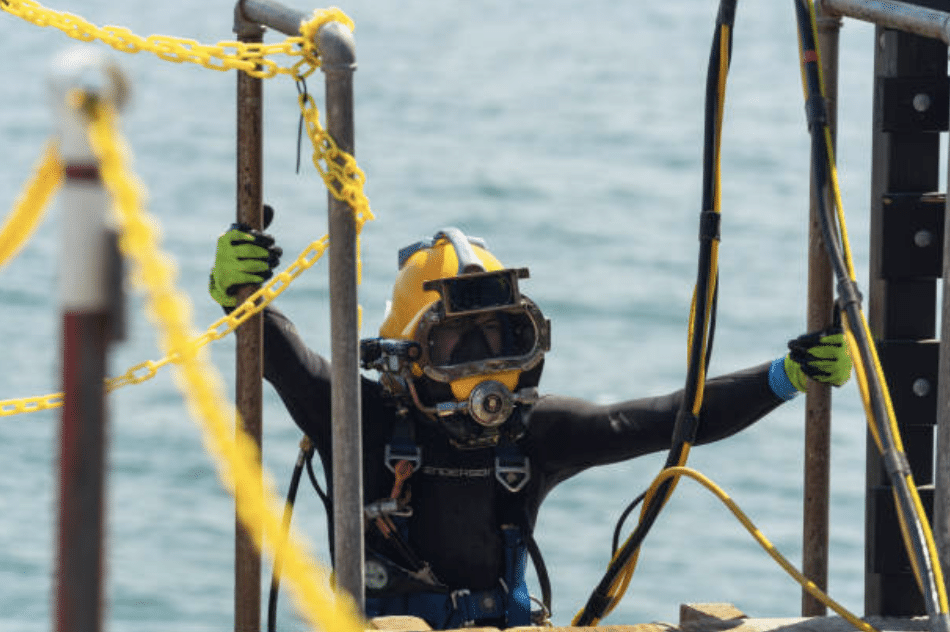 Screenshot 2026-03-15 at 124702 - N9BO℠ | Global Underwater Services Ltd A commercial diver wearing a yellow helmet, black wetsuit, and neon gloves holds onto a metal frame with safety chains near water, preparing for an underwater task.