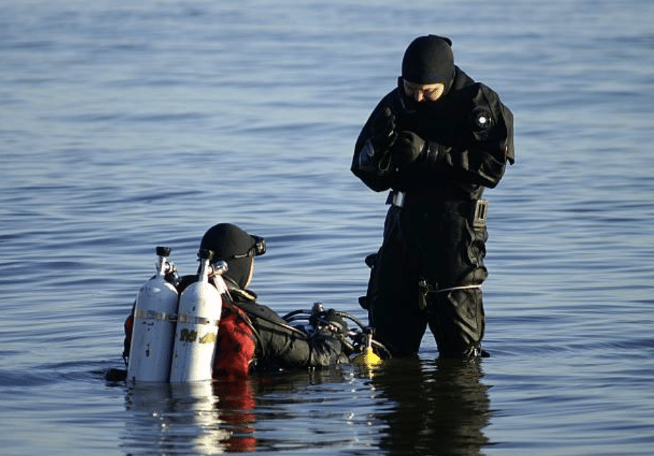 Screenshot 2026-03-15 at 124751 - N9BO℠ | Global Underwater Services Ltd Two scuba divers in black wetsuits stand in calm water, one fully equipped with oxygen cylinders and sitting in the water, the other standing beside, both preparing for a dive.