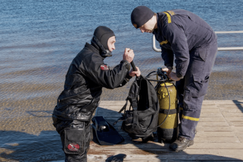 Screenshot 2026-03-15 at 125334 - N9BO℠ | Global Underwater Services Ltd Two people in wetsuits and beanies are preparing scuba diving equipment on a wooden jetty by the water, with one kneeling and the other standing while handling a yellow oxygen cylinder.