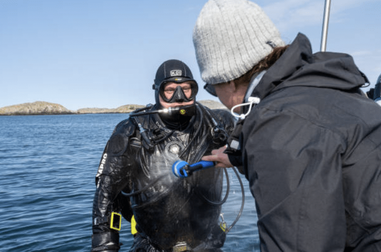 Screenshot 2026-03-15 at 125349 - N9BO℠ | Global Underwater Services Ltd A person in scuba diving gear stands in the water near a boat while another person sprays water on the diver's face with a hose. Rocky islands are visible in the background.