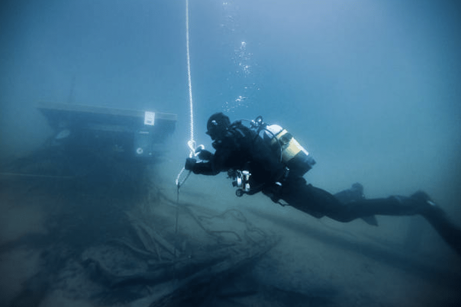 Screenshot 2026-03-15 at 130501 - N9BO℠ | Global Underwater Services Ltd A scuba diver in full kit holds onto a rope underwater, swimming near submerged debris with a structure visible in the background.