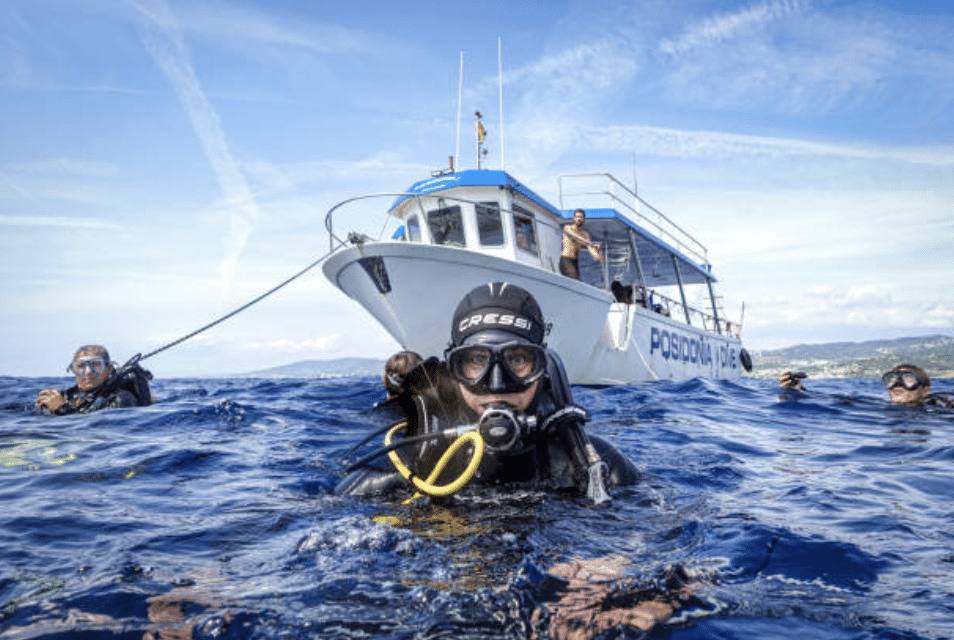 Screenshot 2026-03-15 at 130538 - N9BO℠ | Global Underwater Services Ltd A group of scuba divers floats in the sea near a boat labelled POSIDON DIVE. One diver is close to the camera, wearing a mask and holding a regulator, whilst others and the boat are visible in the background.