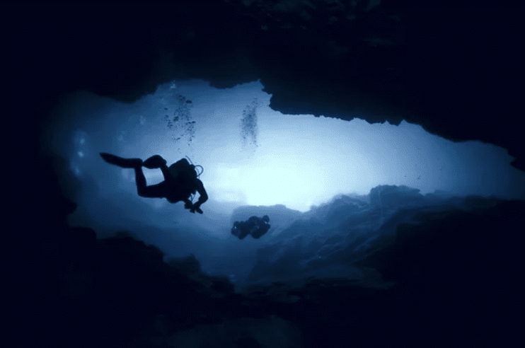 Screenshot 2026-03-15 at 131402 - N9BO℠ | Global Underwater Services Ltd Two scuba divers explore an underwater cave, silhouetted against the blue light coming from the cave entrance, with rocky walls surrounding them.