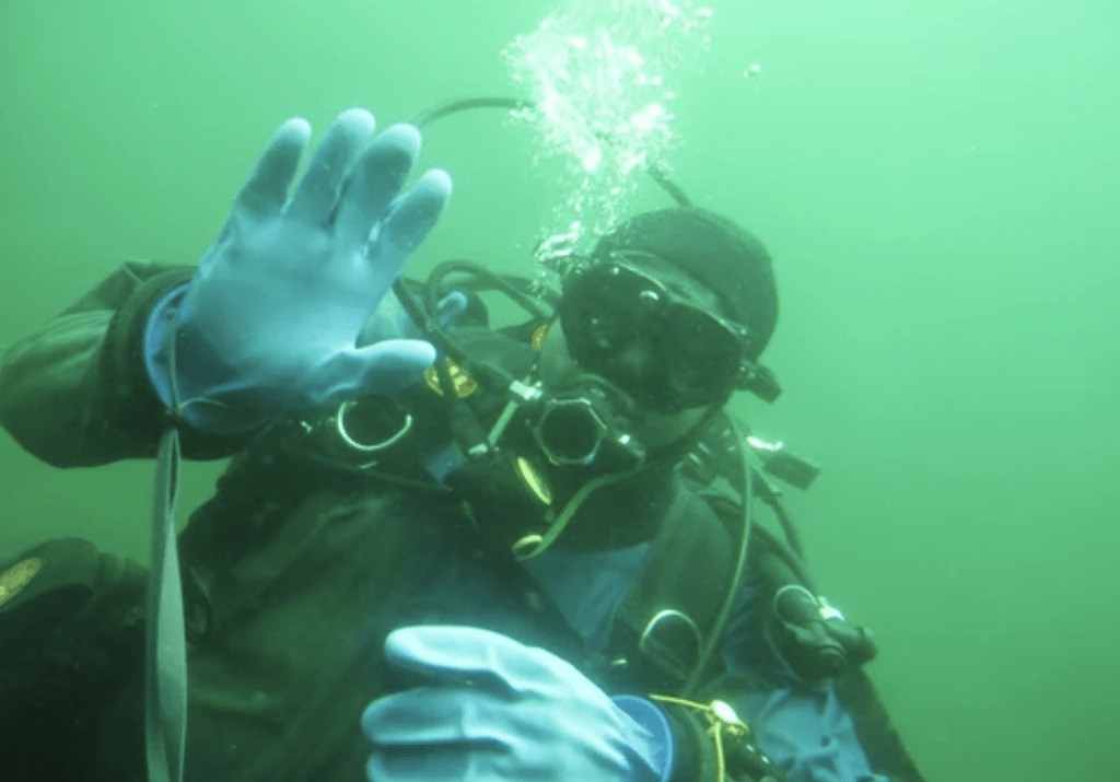 Screenshot 2026-03-15 at 131420 - N9BO℠ | Global Underwater Services Ltd A scuba diver wearing blue gloves, a black mask, and scuba gear waves underwater whilst surrounded by green-tinted water and rising air bubbles.