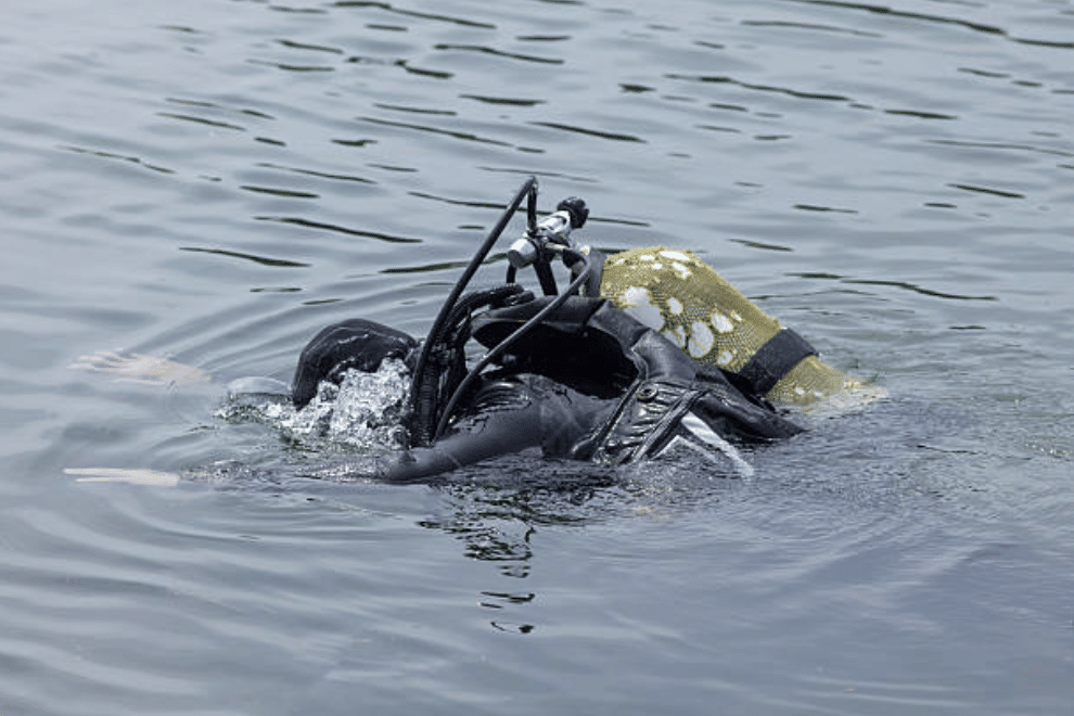 Screenshot 2026-03-15 at 132122 - N9BO℠ | Global Underwater Services Ltd A scuba diver in black kit and a camouflage oxygen cylinder is partially submerged, swimming face-down in calm water.