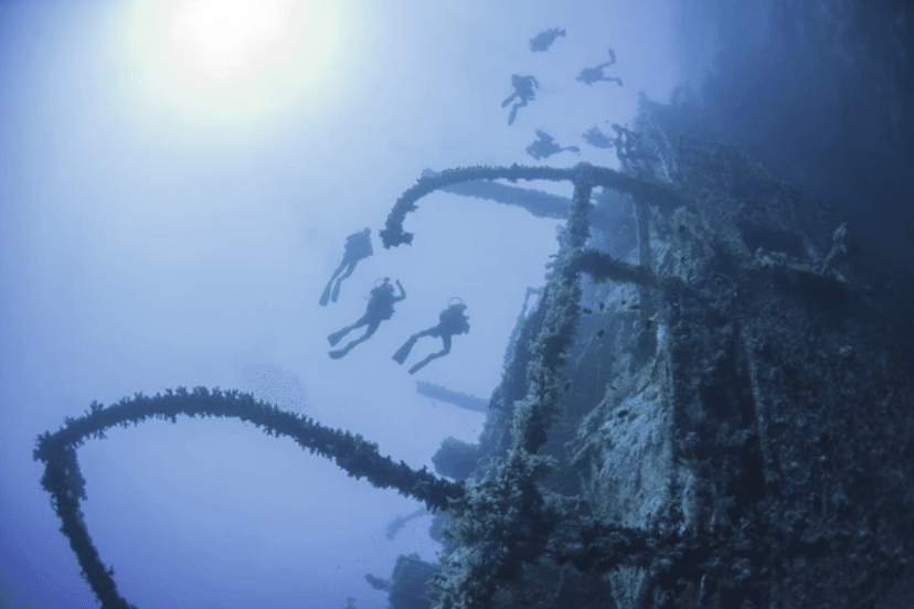 Scuba divers explore a sunken shipwreck deep underwater, surrounded by blue light filtering from the sun above and marine growth covering the ship’s metal structure.