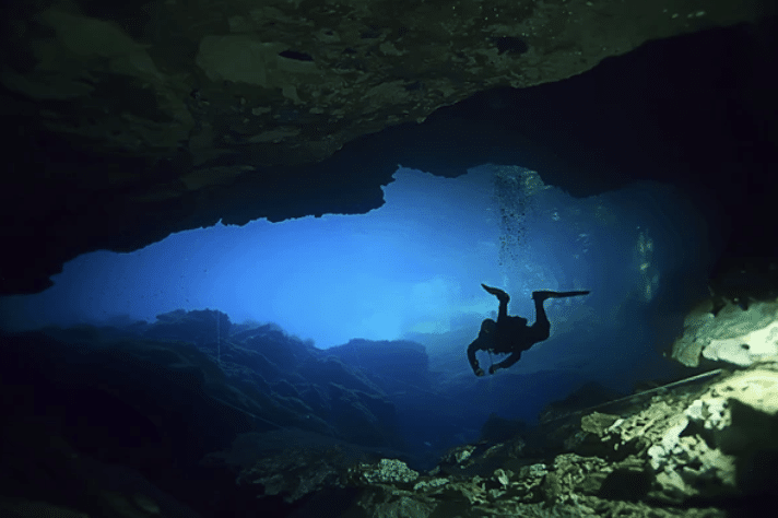 A scuba diver swims through an underwater cave, surrounded by rocky walls and illuminated by blue light from the cave entrance above.
