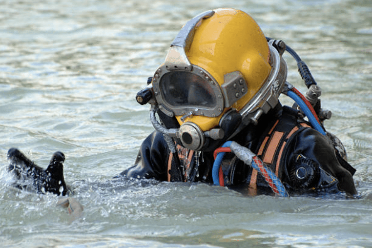 A diver wearing a yellow helmet and a full-face mask suit is partially submerged in water, with gloved hands visible above the surface and air hoses connected to the helmet.