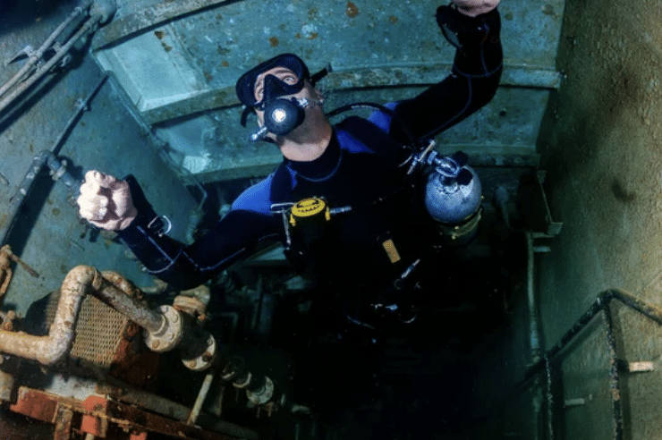 Screenshot 2026-03-15 at 140837 - N9BO℠ | Global Underwater Services Ltd A scuba diver in full kit explores the interior of a sunken, rusted shipwreck, surrounded by pipes and corroded metal structures underwater.