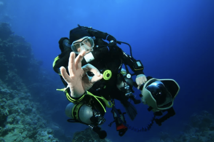 Screenshot 2026-03-15 at 141200 - N9BO℠ | Global Underwater Services Ltd A scuba diver underwater gives an OK hand signal, wearing full diving kit and holding an underwater camera, with a deep blue ocean and coral reef in the background.