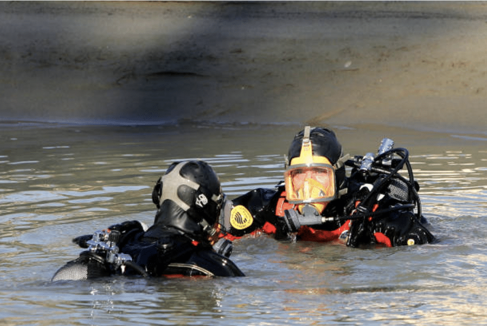 Screenshot 2026-03-15 at 143000 - N9BO℠ | Global Underwater Services Ltd Two divers wearing full kit and helmets are standing in shallow, murky water, facing each other and appearing to communicate, with a sandy embankment visible in the background.