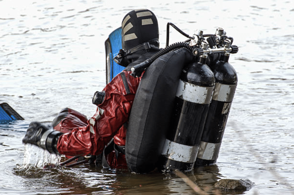 Screenshot 2026-03-15 at 143016 - N9BO℠ | Global Underwater Services Ltd A scuba diver wearing a red drysuit and carrying twin black-and-white air cylinders sits in shallow water, preparing for a dive. The diver's blue fins and full kit are visible.