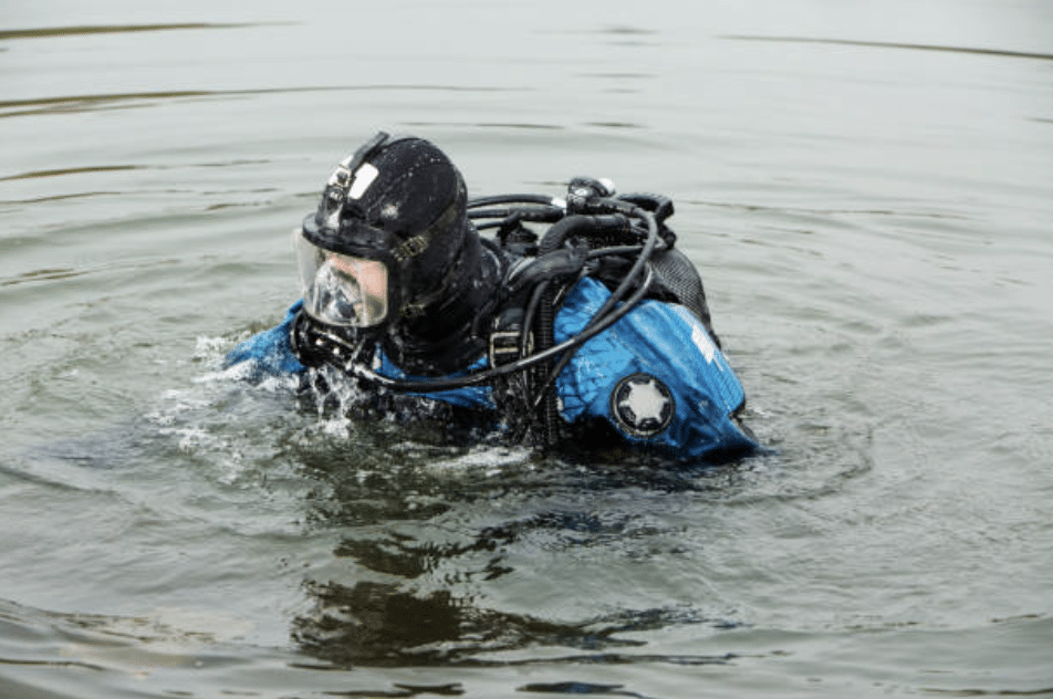 A scuba diver in a black wetsuit and blue diving gear floats at the surface of calm water, with water droplets on their mask and suit.