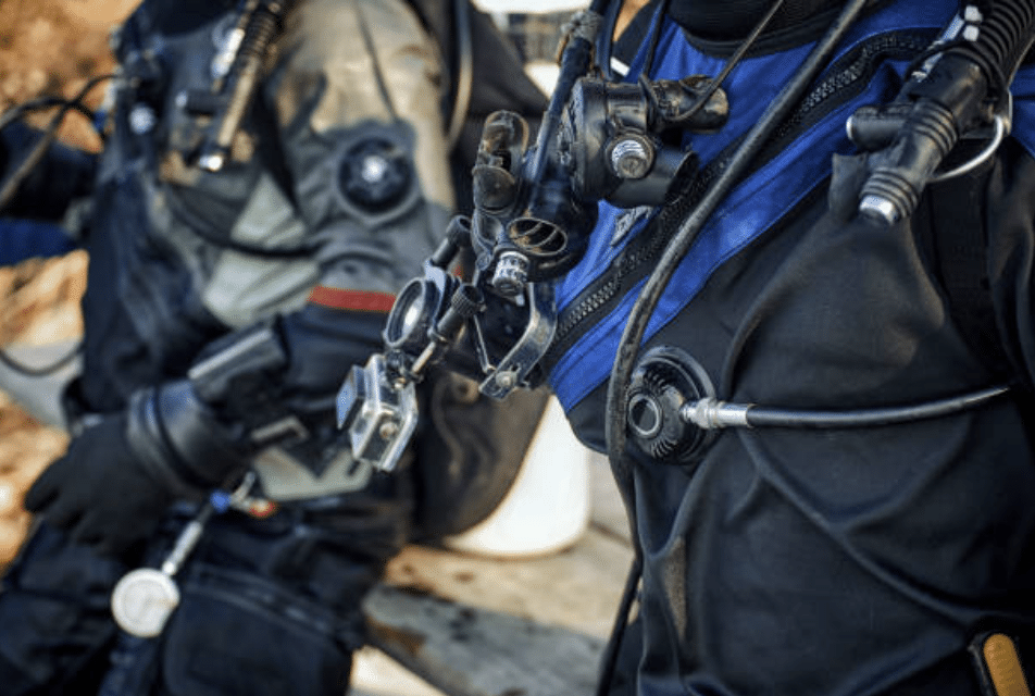 Close-up of two scuba divers in full kit, focusing on their wetsuits, oxygen cylinders, hoses, and breathing apparatus as they prepare for a dive.