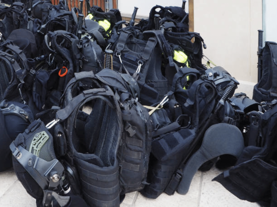 A pile of black tactical vests and body armour lies on the ground, with various straps, clips, and equipment attached, arranged closely together.