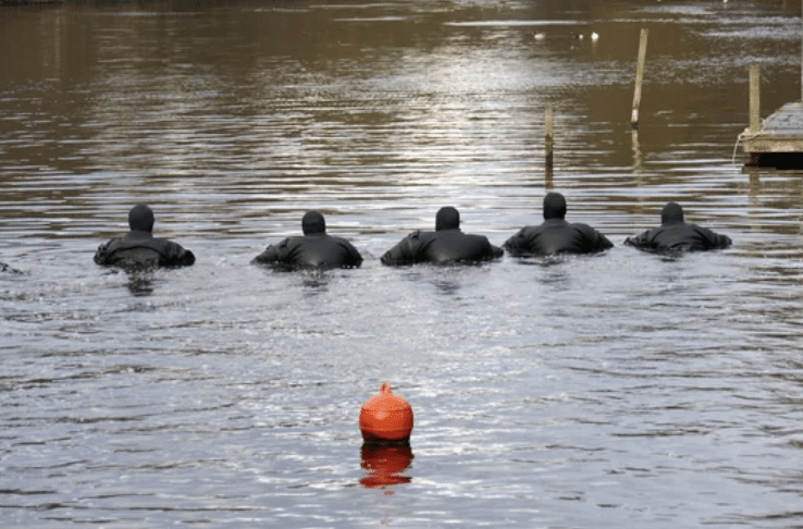 Screenshot 2026-03-15 at 143657 - N9BO℠ | Global Underwater Services Ltd Five people in black wetsuits swim in a line across a body of water, facing away from the camera. A single orange buoy floats in the foreground. The water appears calm with wooden poles in the background.