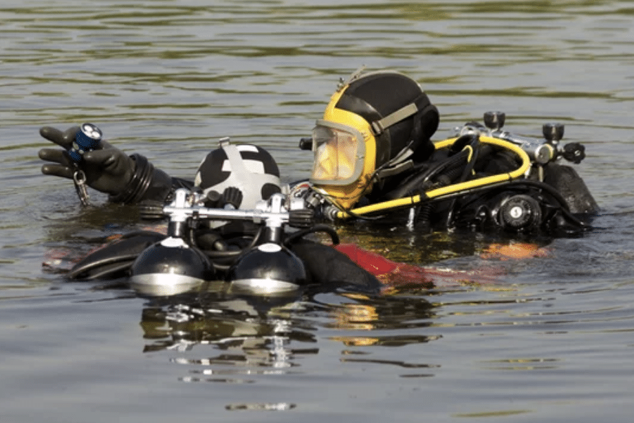 Screenshot 2026-03-15 at 144056 - N9BO℠ | Global Underwater Services Ltd Two scuba divers wearing full-face masks and wetsuits are floating on the surface of the water. One diver points ahead whilst the other faces away, with cylinders and diving equipment visible.