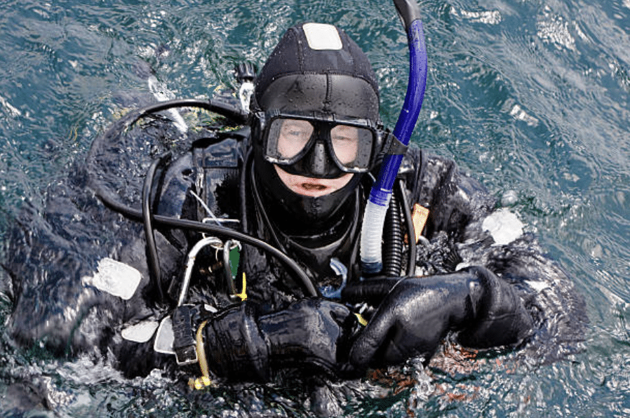 A scuba diver in a black wetsuit, mask, and gloves floats in the water, holding equipment and looking up towards the camera.