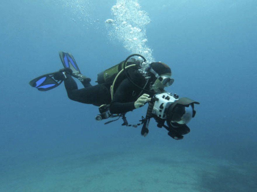 A scuba diver in full kit is underwater, holding a camera in both hands and taking photos or video. The water is clear and blue, with the sea floor faintly visible below.