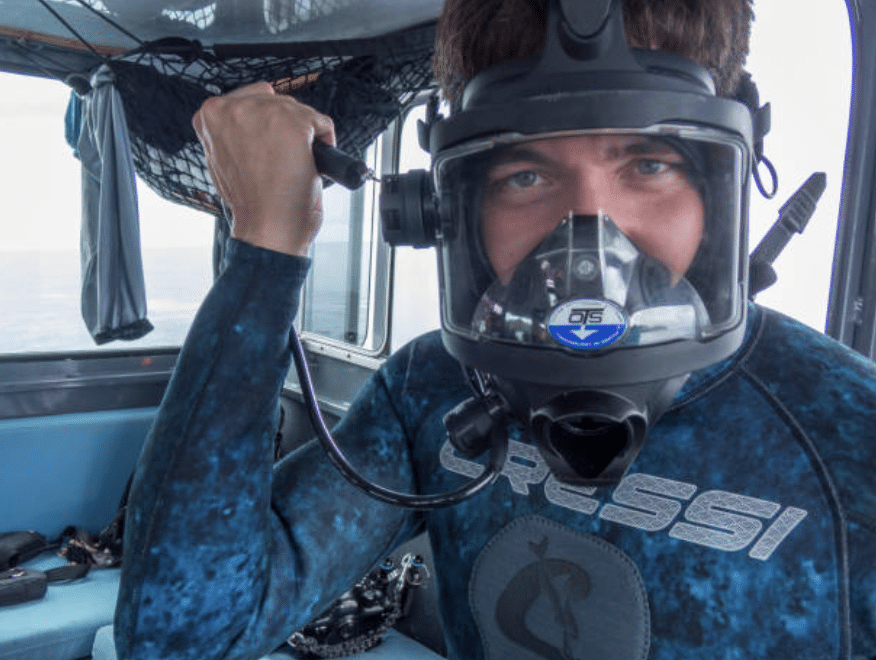 Screenshot 2026-03-15 at 154832 - N9BO℠ | Global Underwater Services Ltd A person in a blue wetsuit and full-face diving mask prepares for a dive inside a boat, holding part of their mask and looking at the camera. Diving equipment is visible in the background.