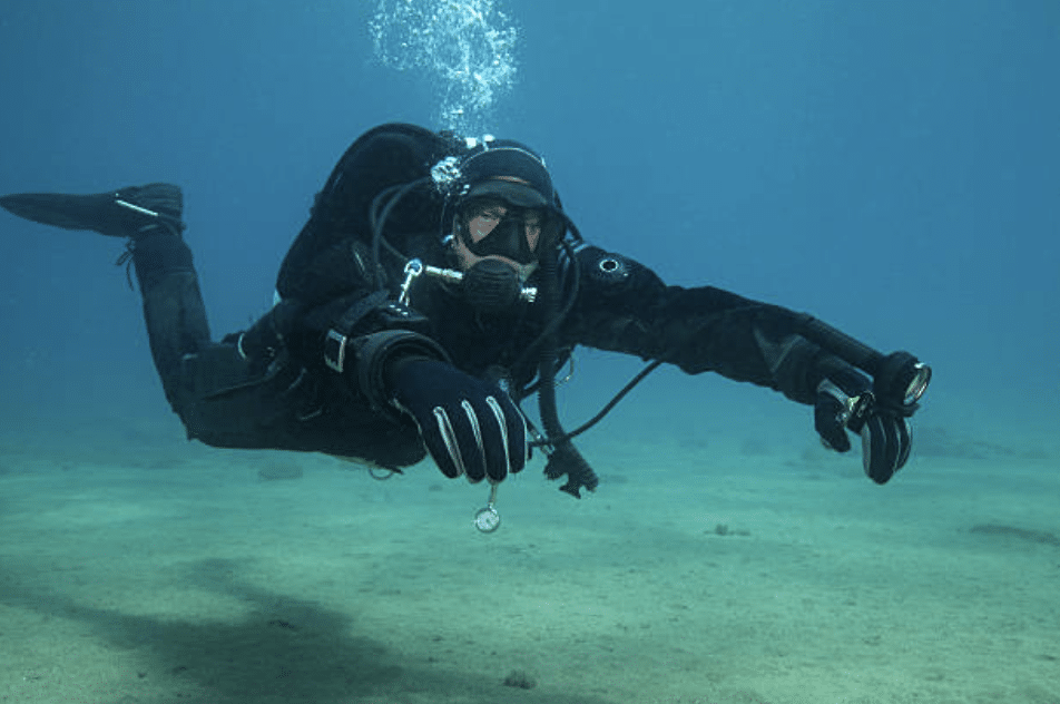 A scuba diver in a black wetsuit and gloves swims underwater, holding a compass and other diving instruments, with bubbles rising above them and the sandy sea floor visible below.