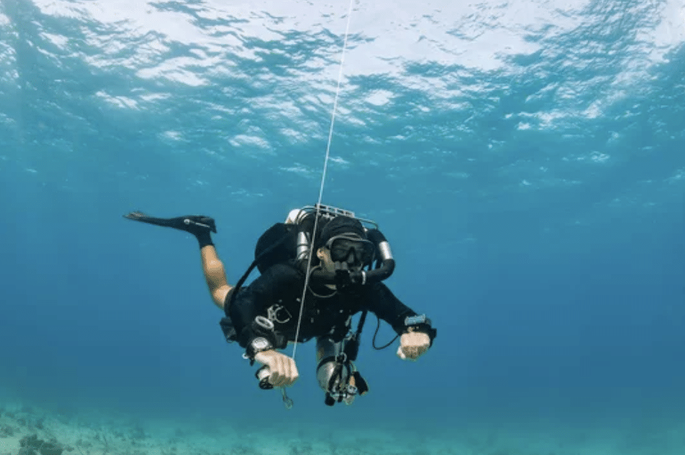 A scuba diver in full kit is underwater, holding onto a guide line descending from the surface. Sunlight filters through the clear blue water, and the seabed is visible below.