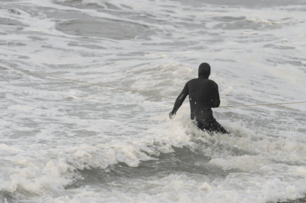 Screenshot 2026-03-15 at 170021 - N9BO℠ | Global Underwater Services Ltd A person wearing a dark, hooded outfit stands in rough sea waves, holding onto a rope that stretches across the water.