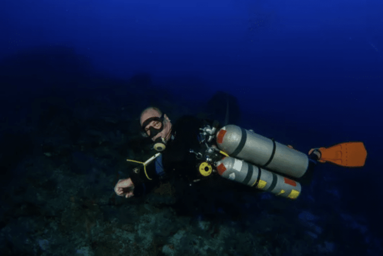 A scuba diver wearing a black wetsuit and orange fins swims underwater with multiple air cylinders attached to their back in deep blue water.