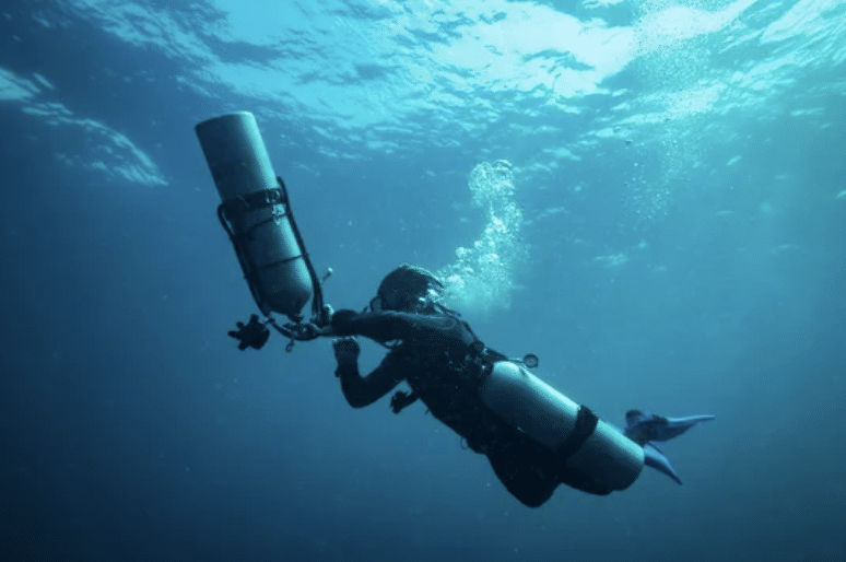 Screenshot 2026-03-15 at 171517 - N9BO℠ | Global Underwater Services Ltd A scuba diver swims underwater, equipped with multiple air tanks and diving gear, with sunlight streaming down through the blue sea above. Bubbles rise from the diver’s mask as they explore the deep water.