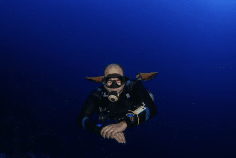 Screenshot 2026-03-15 at 171537 - N9BO℠ | Global Underwater Services Ltd A scuba diver wearing a wetsuit, mask, and cylinder swims underwater in deep blue ocean water, with their hands folded in front and no visible marine life around.