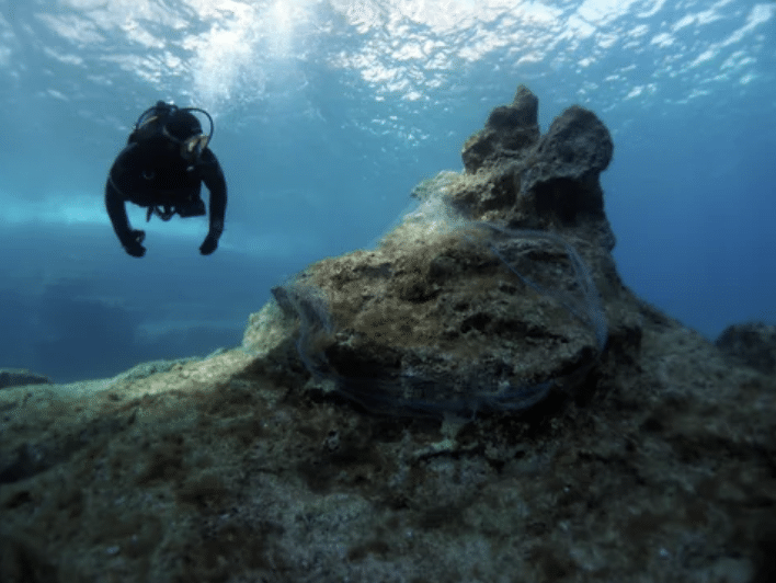 Screenshot 2026-03-15 at 172615 - N9BO℠ | Global Underwater Services Ltd A scuba diver swims underwater near a rocky formation covered with fishing nets, surrounded by clear blue water and sunlight filtering from above.