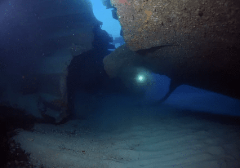 Screenshot 2026-03-15 at 174241 - N9BO℠ | Global Underwater Services Ltd A diver with a torch explores the interior of a sunken shipwreck underwater, surrounded by sandy seabed and blue light filtering through gaps in the wreck's structure.
