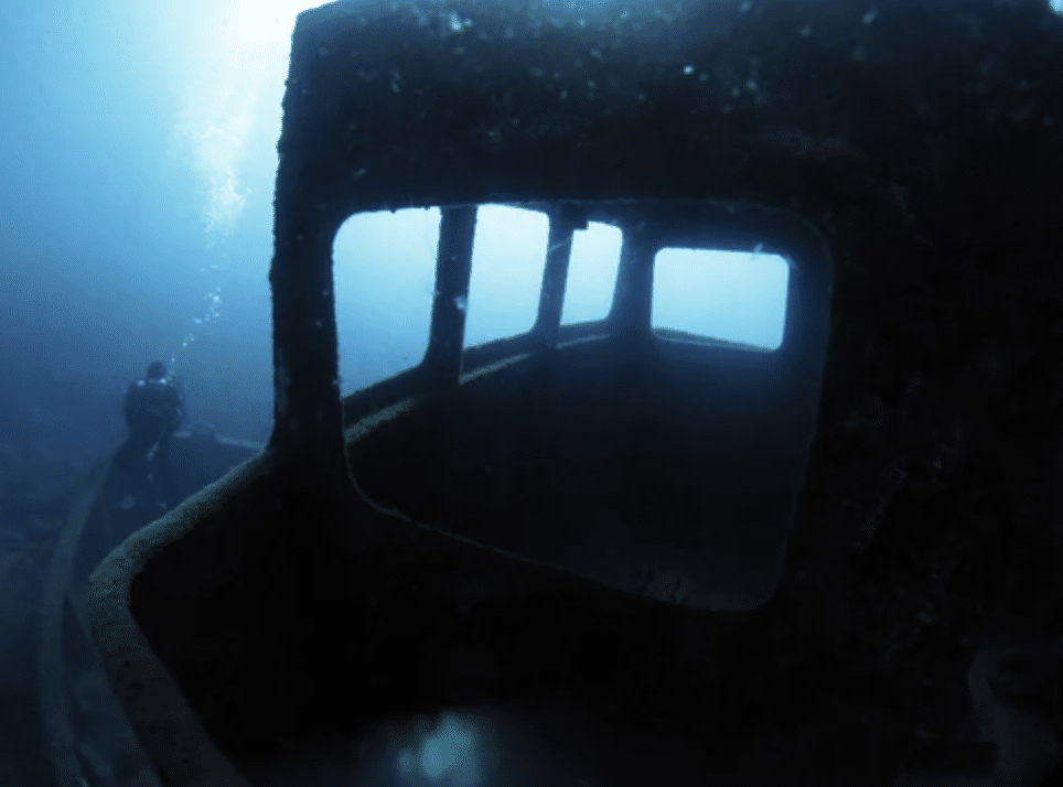 Screenshot 2026-03-15 at 174253 - N9BO℠ | Global Underwater Services Ltd A scuba diver explores a sunken shipwreck underwater, viewed through the rusted window frame of the ship's cabin, surrounded by deep blue water.