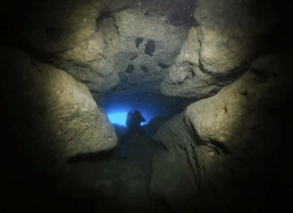 Screenshot 2026-03-15 at 174301 - N9BO℠ | Global Underwater Services Ltd A person scuba-dives through an underwater cave or tunnel, silhouetted against a distant blue light shining from the cave’s exit, surrounded by large rocky walls.
