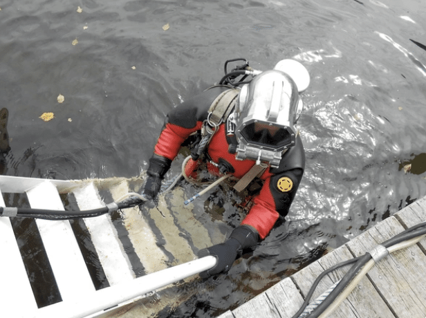 Screenshot 2026-03-15 at 182656 - N9BO℠ | Global Underwater Services Ltd A diver in a red and black drysuit with a helmet climbs a white ladder from the water onto a wooden jetty, holding onto a handrail with cables attached to their suit.