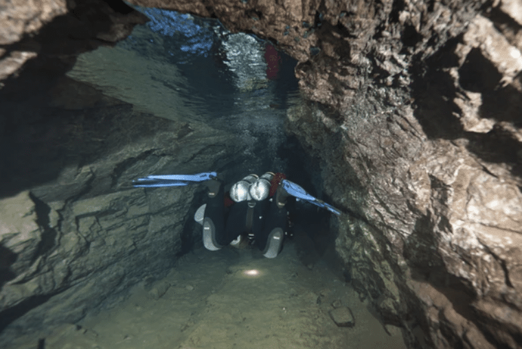 Screenshot 2026-03-15 at 182944 - N9BO℠ | Global Underwater Services Ltd A scuba diver with blue fins and a headtorch swims through a narrow, rocky underwater cave passage, illuminating the dark tunnel ahead.