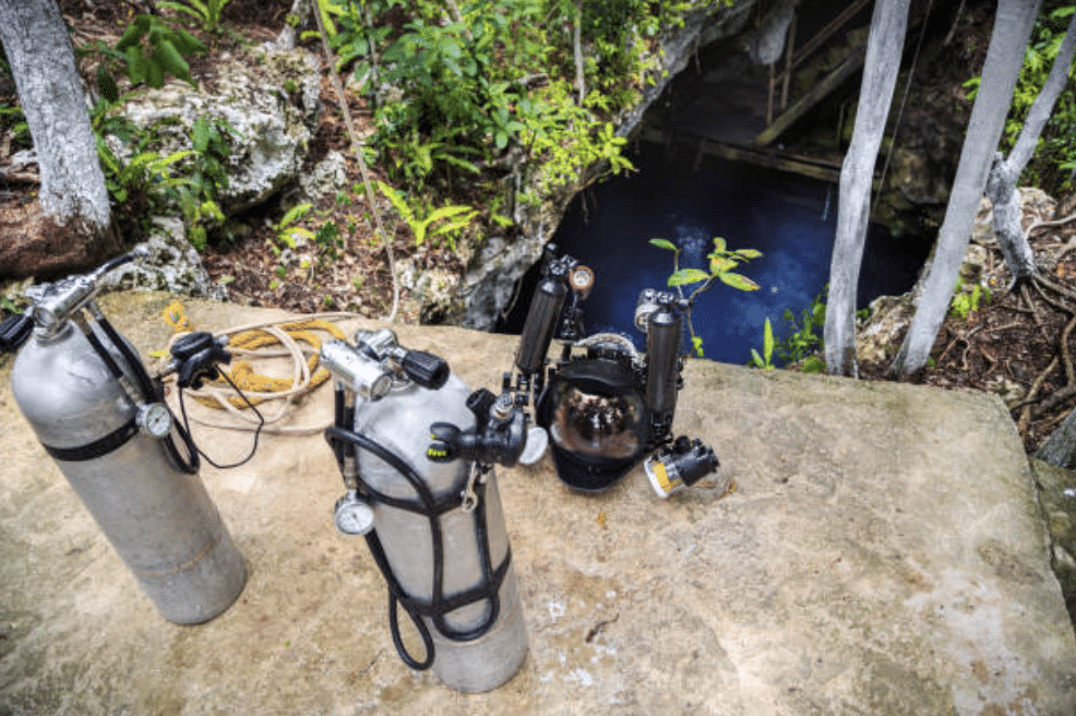Screenshot 2026-03-15 at 183034 - N9BO℠ | Global Underwater Services Ltd Several scuba cylinders, diving kit, and an underwater camera are placed on a stone surface near the entrance of a water-filled cave surrounded by lush greenery.