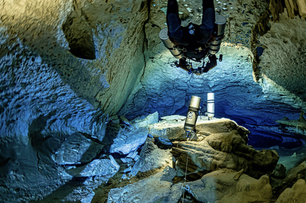Screenshot 2026-03-15 at 183044 - N9BO℠ | Global Underwater Services Ltd A scuba diver with multiple cylinders explores an underwater cave filled with rocks and illuminated by blue light.