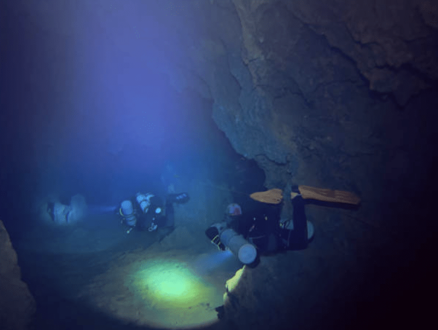 Two scuba divers with torches explore a dark underwater cave, illuminating the rocky walls and sandy floor as they navigate through the confined, shadowy passage.