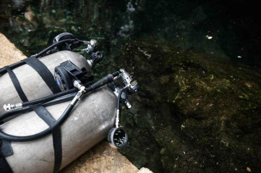 Screenshot 2026-03-15 at 184200 - N9BO℠ | Global Underwater Services Ltd Two scuba diving air cylinders with attached gauges and hoses rest on a concrete surface near the edge of water, ready for use in a dive.