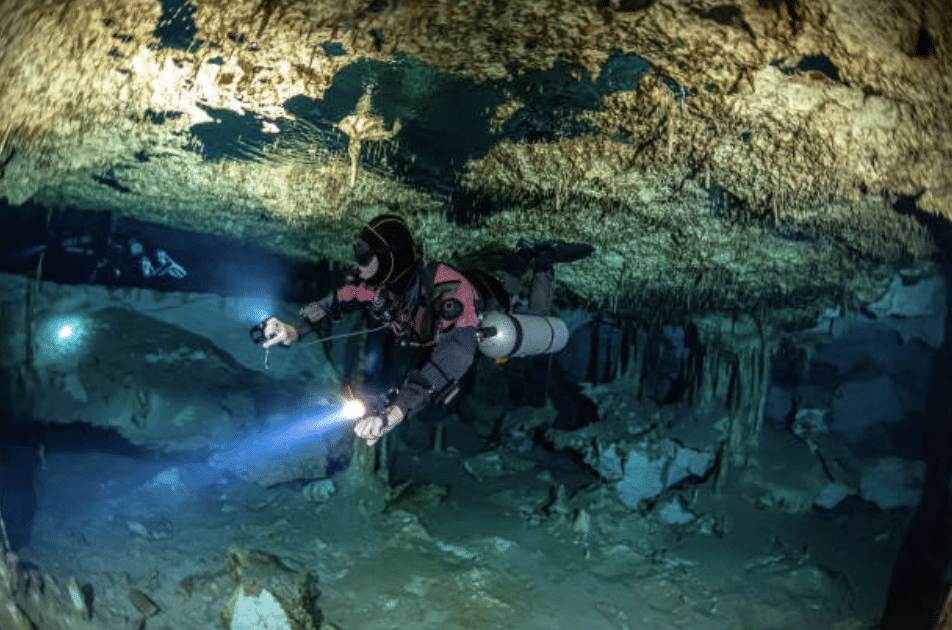 Screenshot 2026-03-15 at 184210 - N9BO℠ | Global Underwater Services Ltd A scuba diver in a wetsuit explores an underwater cave, shining a torch on the rocky ceiling surrounded by stalactites and submerged rock formations.