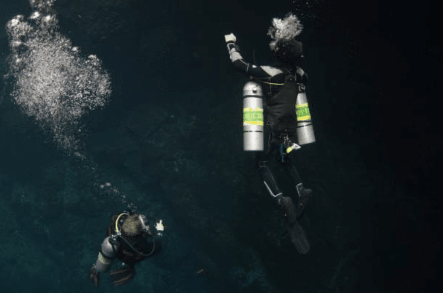 Screenshot 2026-03-15 at 184223 - N9BO℠ | Global Underwater Services Ltd Two scuba divers with double oxygen cylinders swim underwater in dark, clear water. Bubbles rise above them as one diver gestures upward, surrounded by a deep blue and black background.