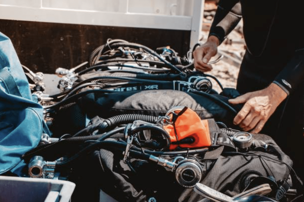 Screenshot 2026-03-15 at 194252 - N9BO℠ | Global Underwater Services Ltd A person in a wetsuit prepares scuba diving equipment, adjusting cylinders and regulators, with various hoses and an orange item among the kit.